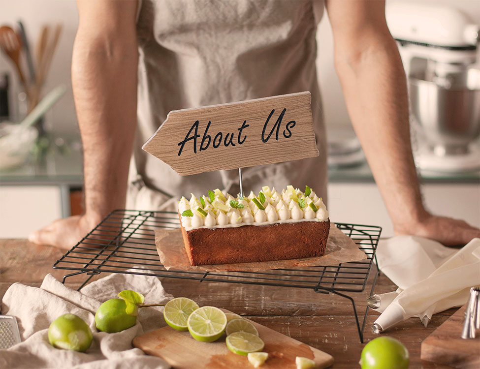 Baker standing in front of decorated cake with sign displaying About Us